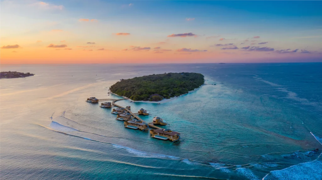 Aerial view of a private island resort surrounded by turquoise waters at sunset, featuring overwater villas and lush greenery—showcasing eco-luxury and climate-resilient island living.
